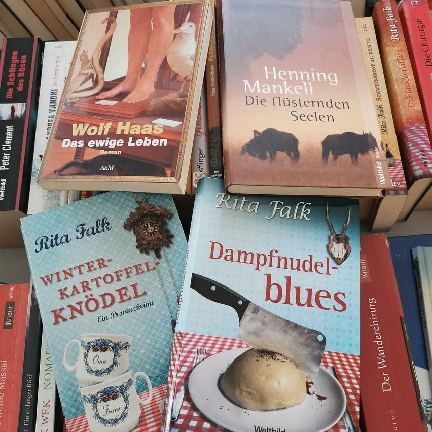 Zusammengestapelte Bücher umfassen Titel wie Wolf Haas Das ewig Leben und Henning Mankell Die flüsternden Seelen. Ein Buch mit einem Messer und Essen auf dem Cover heißt Dampfnudel blues.