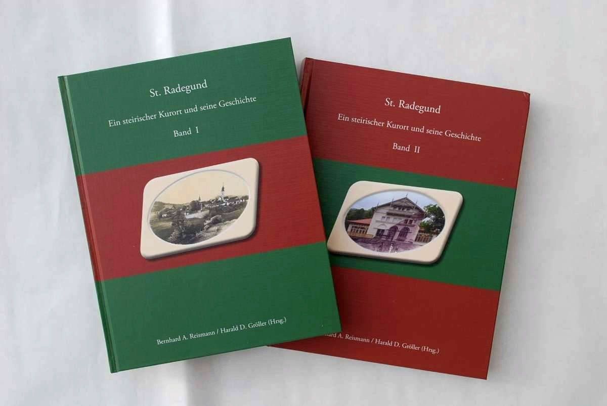 Two books, titled 'St. Radegund: A Styrian Spa Town and Its History,' are displayed. Both are green and red with white borders. Volume 1 features an old image of a landscape, while Volume 2 shows an image of a building.