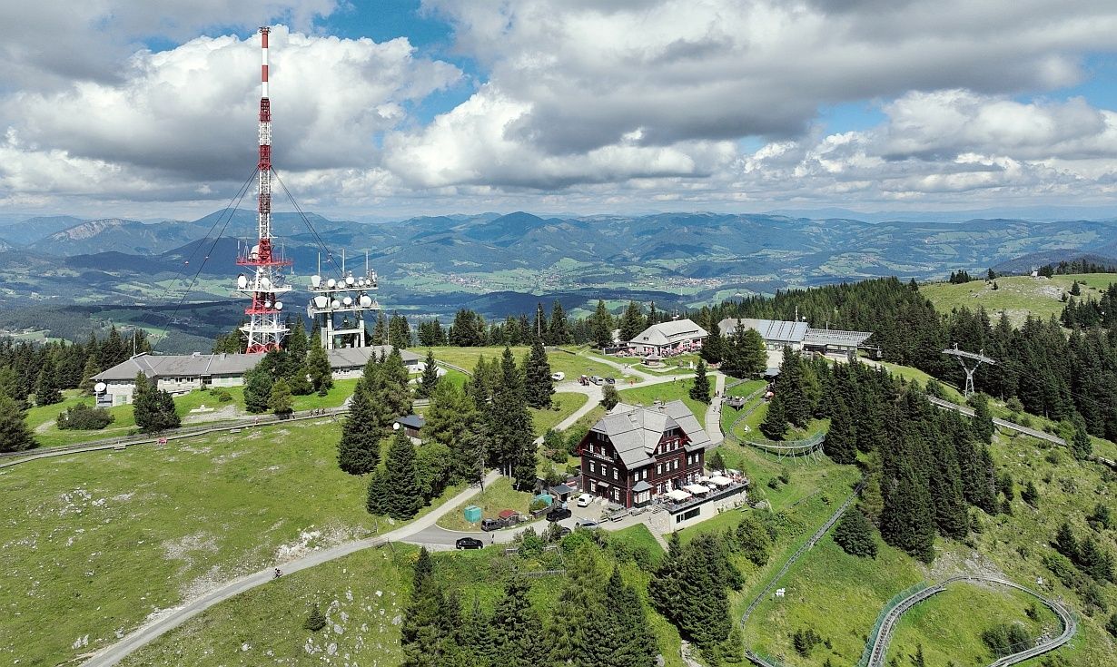 Luftaufnahme eines Berggipfels mit einem Turm, Häusern und üppigem Grün. Im Hintergrund Berge und ein bewölkter Himmel.