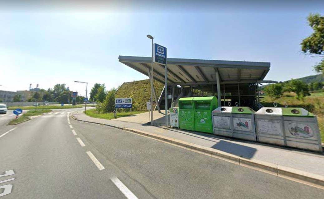 Ein Parkplatz mit einem überdachten Unterstand, einem Schild mit der Aufschrift P+R und mehreren grünen Mülleimern. Die Straße ist leer mit weißen Markierungen.
