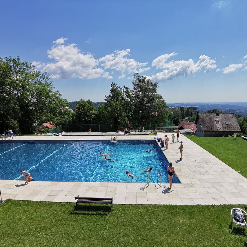 Ein Blick von oben auf ein Freibad mit mehreren schwimmenden Personen und einigen am Rand. Bäume und ein Haus sind im Hintergrund.