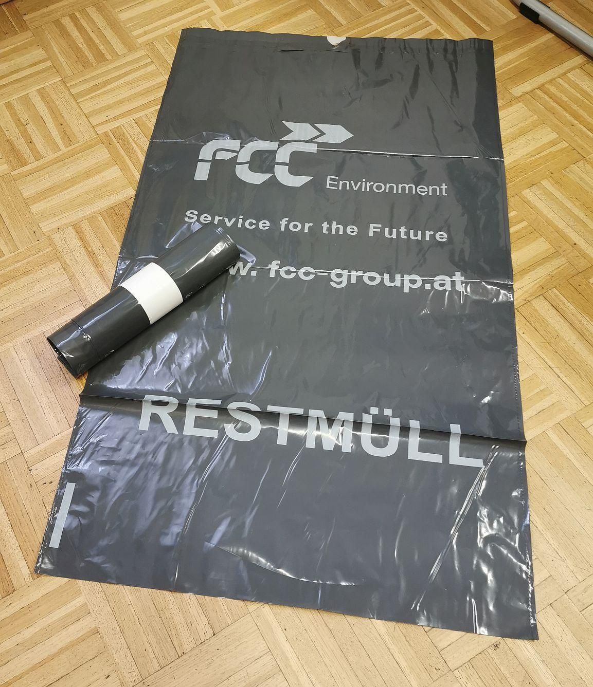 Ein schwarzer Plastiksack mit der Aufschrift 'FCC Environment' und 'Service for the Future' liegt auf einem Holzboden. 'RESTMULL' ist weiß aufgedruckt.