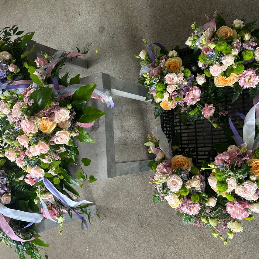 Zwei Blumenarrangements mit rosa, gelben und blauen Blumen sind auf einem Betonboden ausgestellt. Jedes Arrangement ist mit lila und weißen Bändern gebunden.