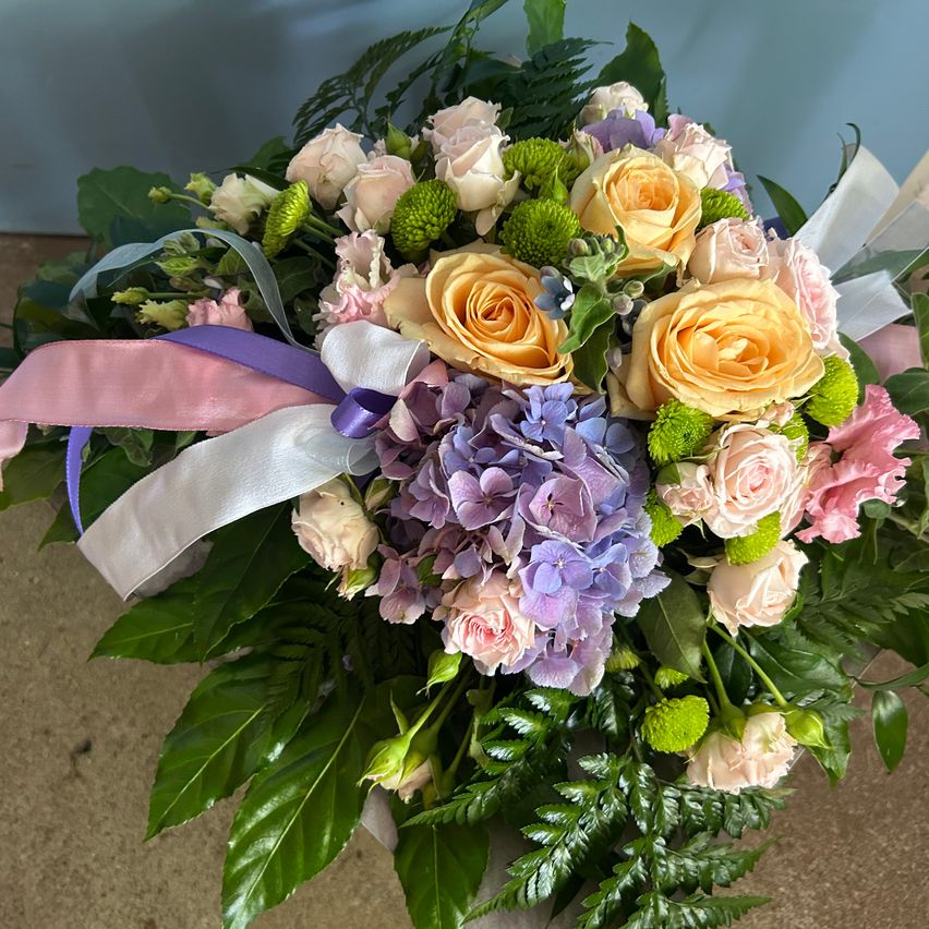 Ein farbenfrohes Blumenarrangement besteht aus einer Mischung aus weißen, rosa und gelben Rosen sowie lila und blauen Hortensien. Verziert mit grünen Blättern, ist es mit weißen und lilafarbenen Bändern gebunden.