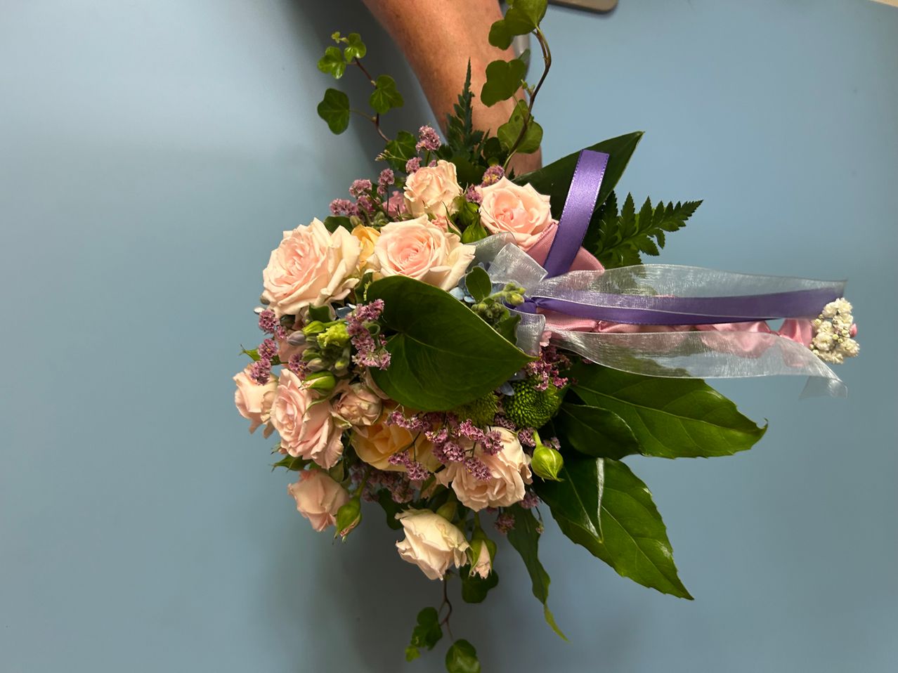 Ein Strauß aus hellrosa und weißen Rosen mit grünen Blättern und einem lila Band wird von einer Hand vor einem blauen Hintergrund gehalten.