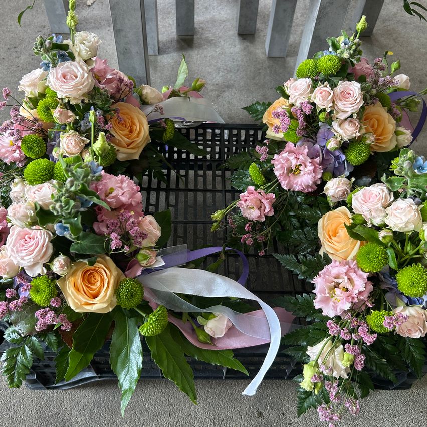 Drei Blumengestecke in Form von Kränzen, jedes gefüllt mit einer Vielzahl von Blumen, darunter Rosen, Hortensien und Grünpflanzen, auf einem schwarzen Korb platziert.