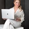 Eine Frau in weißer Kleidung mit professionellem Auftreten sitzt auf einer weißen Bank und hält einen Apple Laptop.