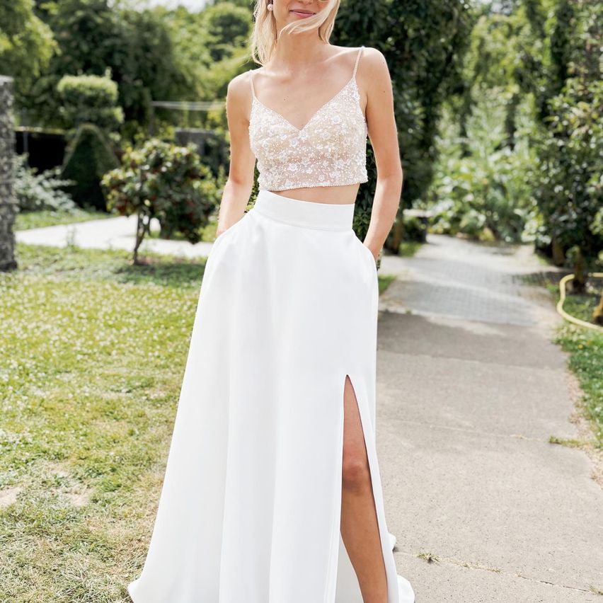 Eine Frau in einem weißen Kleid mit hohem Schlitz steht auf einem Betonweg in einem Garten.
