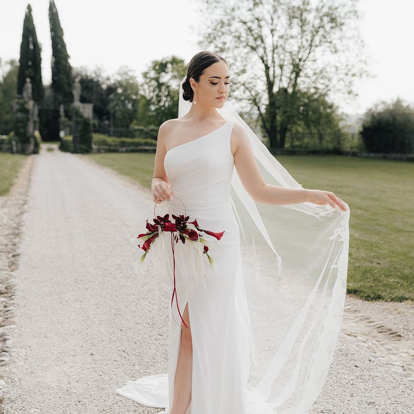 Eine Braut in einem weißen Kleid mit Schleier geht einen Kiesweg entlang und hält einen Strauß roter Blumen. Im Hintergrund sind Bäume und Grün zu sehen.