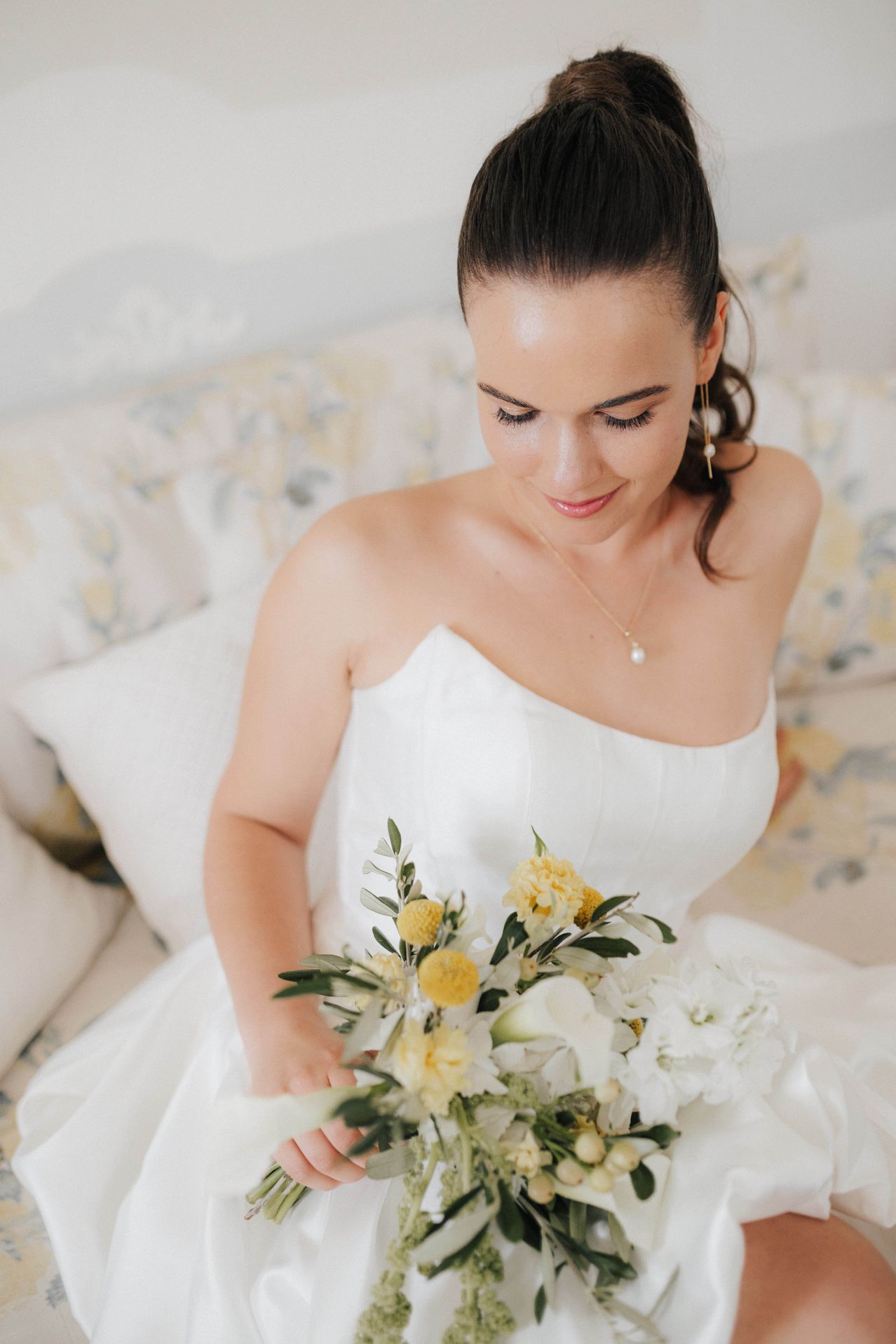 Eine Braut in einem weißen Kleid sitzt auf einer Blumencouch und hält einen Strauß gelber und weißer Blumen. Sie lächelt und blickt nach unten.