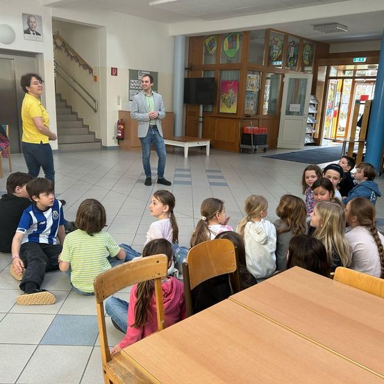 Ein Mann und eine Frau stehen in einem Klassenzimmer mit Schülern, die um sie herum sitzen und zuhören. Das Klassenzimmer hat blaue Säulen, eine Treppe und einen Fernseher auf einem Ständer.
