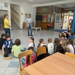 Ein Mann und eine Frau stehen in einem Klassenzimmer mit Schülern, die um sie herum sitzen und zuhören. Das Klassenzimmer hat blaue Säulen, eine Treppe und einen Fernseher auf einem Ständer.