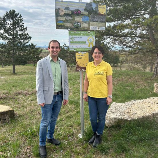 Zwei Personen stehen neben einem Wegweiser in einem Grasfeld mit Schildern und Bäumen im Hintergrund.