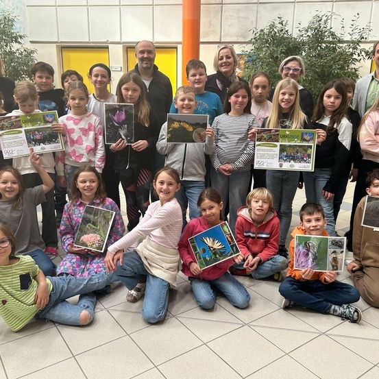 Eine Gruppe von Kindern und Lehrern posiert für ein Foto und hält Urkunden in den Händen. Einige Kinder sitzen auf dem Boden, andere stehen. Sie befinden sich in einem Schulkorridor.