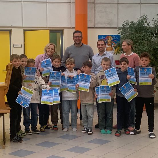 Eine Gruppe von Kindern in einem Schulflur, alle mit Urkunden in der Hand, posiert für ein Foto. Die Kinder stehen vor einem Mann und einer Frau mit lächelnden Gesichtern.