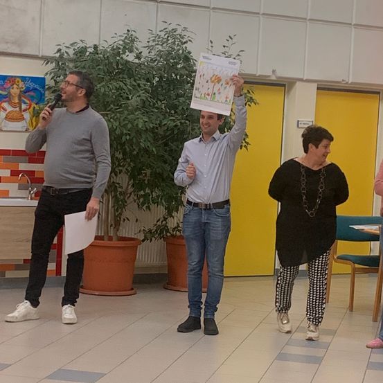 Drei Personen stehen in einem Raum. Der Mann auf der linken Seite hält ein Mikrofon, der Mann in der Mitte hält ein Papier und die Frau auf der rechten Seite lächelt.