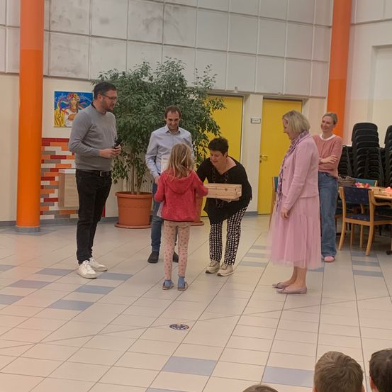 Eine Gruppe von Menschen steht in einem Raum, eine Mädchen erhält eine Auszeichnung. Das Mädchen hält eine Holzkiste, während andere zusehen und herumstehen.