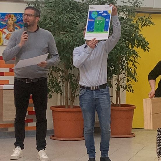 Zwei Männer stehen in einem Raum. Ein Mann hält ein Plakat und der andere ein Mikrofon und ein Stück Papier. Hinter ihnen hält eine Person eine Holzkiste. Der Raum hat eine farbenfrohe Wand und Topfpflanzen.