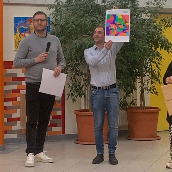 Zwei Männer stehen in einem Raum mit einem farbenfrohen Poster und einem Mikrofon. Der Mann auf der linken Seite hält ein Blatt Papier und ein Mikrofon. Der Mann auf der rechten Seite hält ein farbenfrohes Poster.