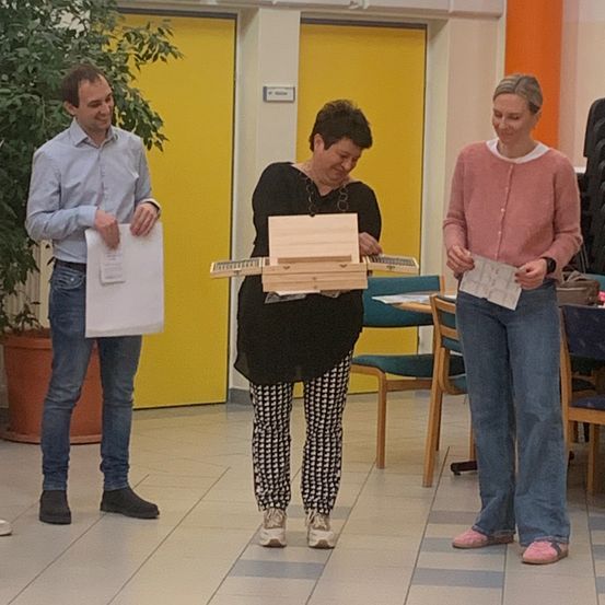 Drei Personen stehen in einem Raum mit gelben Türen. Die Frau in der Mitte hält eine Holzkiste mit einem kleinen Klavier. Der Mann auf der linken Seite hält ein Blatt Papier und die Frau auf der rechten Seite hält ein Blatt Papier und schaut darauf.