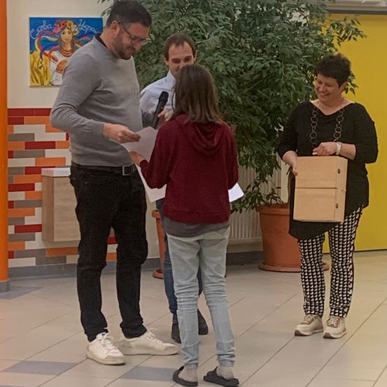 Drei Personen stehen in einem Raum. Ein Mann mit Mikrofon steht neben einer Frau mit Papier, und eine weitere Frau steht hinter ihnen mit einer Holzkiste.