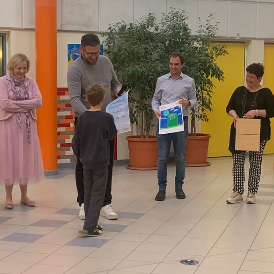 Zwei Männer und zwei Frauen stehen vor einem Jungen, der ein Zertifikat in der Hand hält. Der Junge steht zwischen zwei Männern. Dahinter befinden sich zwei Topfpflanzen.