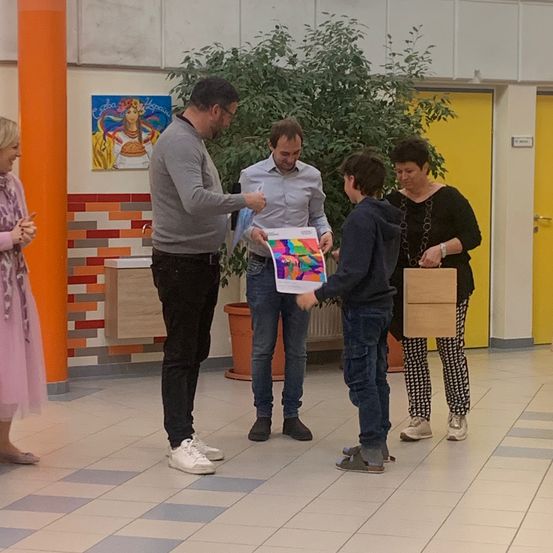 Vier Personen stehen in einem Schulflur, zwei von ihnen halten ein farbenfrohes Poster und zwei von ihnen halten Papiertüten.