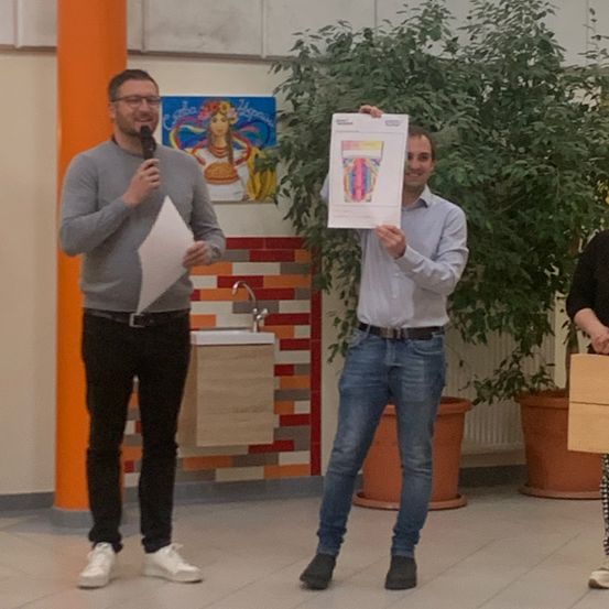 Zwei Männer in einem Raum, einer hält ein Mikrofon und der andere ein Stück Papier. Dahinter befindet sich ein Waschbecken und eine Pflanze. Eine Frau steht rechts.