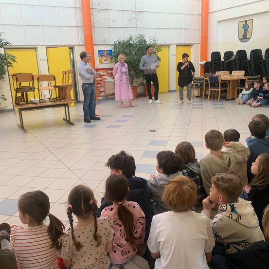 Eine Gruppe von Kindern sitzt im Kreis und beobachtet drei Erwachsene auf der Bühne in einer Schule.