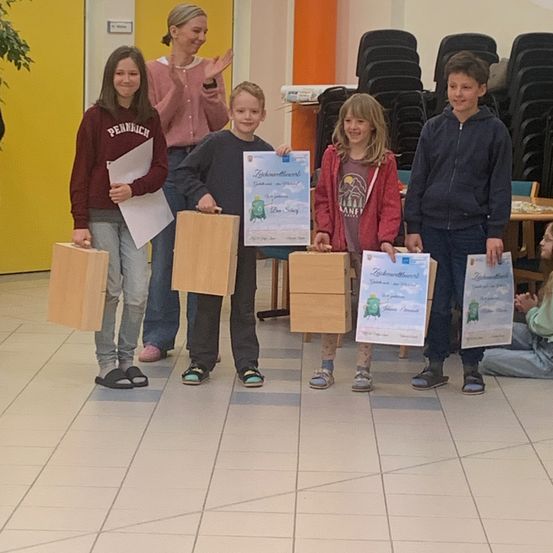 Fünf junge Kinder stehen in einem Raum und halten Urkunden und Holzkisten. Das Mädchen links trägt ein lila Sweatshirt mit dem Wort 'Pennrich'. Eine Frau steht hinter ihnen und klatscht.