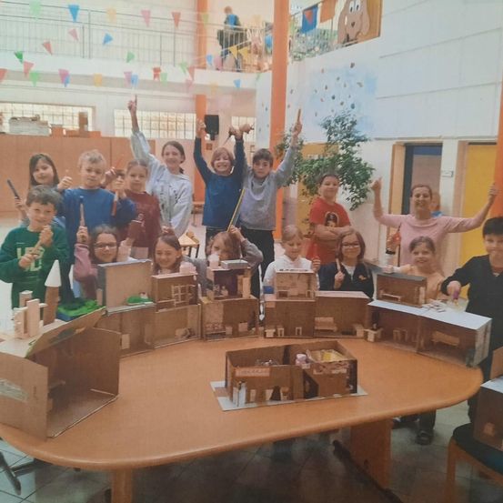 Eine Gruppe von Kindern versammelt sich um einen großen Tisch mit Pappkartons und nimmt wahrscheinlich an einer Bastelaktivität teil. Einige heben ihre Hände, möglicherweise um Fragen zu stellen oder Antworten zu geben. Der Schauplatz scheint ein Klassenzimmer zu sein.