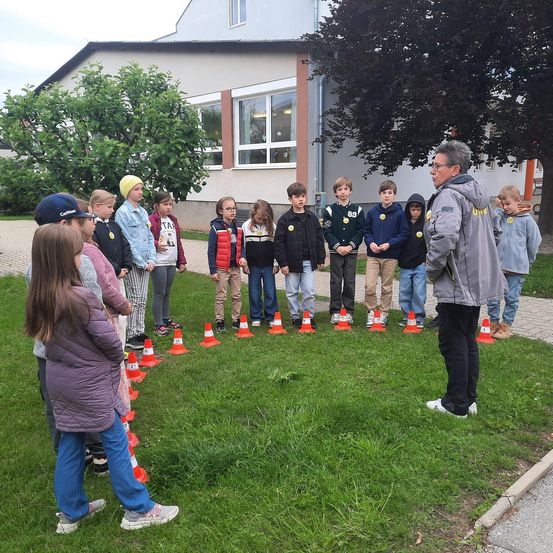 Eine Gruppe von Kindern steht zwischen orangefarbenen Kegeln in einer Reihe, mit einem Erwachsenen, wahrscheinlich einem Lehrer, vor ihnen.