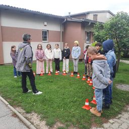 Eine Gruppe von Kindern und einem Erwachsenen steht in einem Kreis aus orangefarbenen Verkehrskegeln vor einem Gebäude.