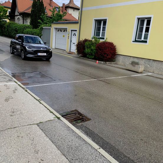 Ein schwarzes Auto fährt auf einer nassen Straße mit einem Abflussdeckel und einem Verkehrskegel in der Nähe eines gelben Gebäudes mit einer weißen Tür.