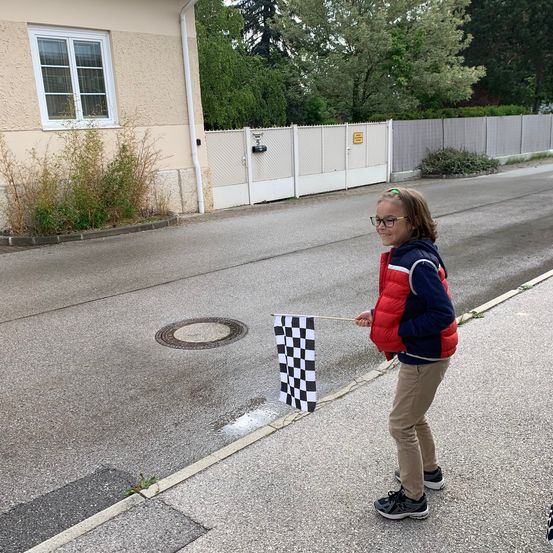 Ein Mädchen in einem roten Weste und Brille hält eine Schachbrettflagge auf einer Straße in der Nähe eines Hauses mit einem weißen Fenster.