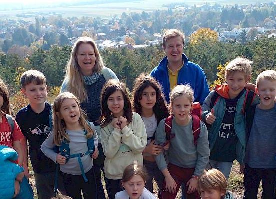 Eine Gruppe von Kindern und zwei Erwachsenen steht auf einem Hügel und lächelt für ein Foto. Sie tragen Rucksäcke und einige halten sich aneinander fest. Im Hintergrund befinden sich Bäume, eine Stadt und ein klarer Himmel.