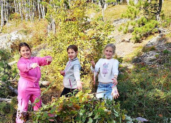 Drei Mädchen stehen in einem Wald, lächeln und halten Stöcke. Das Mädchen auf der linken Seite trägt einen rosa Schlafanzug, während die anderen weiße und graue Jacken tragen.