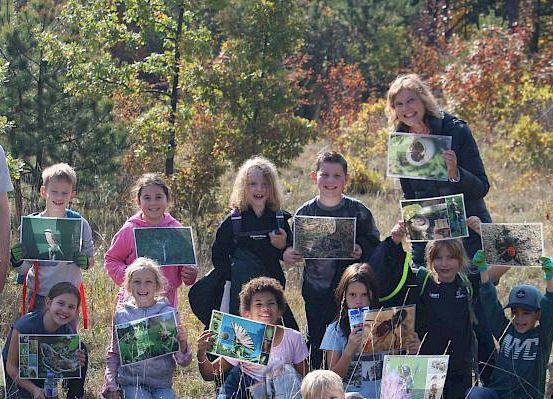Eine Gruppe von Kindern und zwei Erwachsenen posiert auf einem Feld mit Bäumen. Sie halten Fotos von Vögeln und Pflanzen hoch. Die Erwachsenen lächeln.