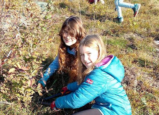 Zwei junge Mädchen bücken sich in einem grasigen Bereich und schneiden mit Gartenschere Äste von einem Busch ab. Ein Mädchen trägt eine blaue Jacke mit Kapuze.