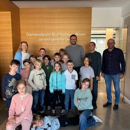 Eine Gruppe von Kindern und Erwachsenen posiert für ein Foto vor einer hölzernen Wand mit Text auf Deutsch. Sie alle sehen fröhlich und aufgeregt aus.