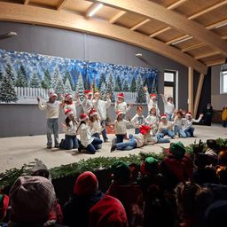 Eine Gruppe von Kindern in festlichen Mützen und Schals tritt auf einer Bühne auf, die mit Weihnachtsbäumen und Grünpflanzen geschmückt ist. Ein Publikum beobachtet von unten.