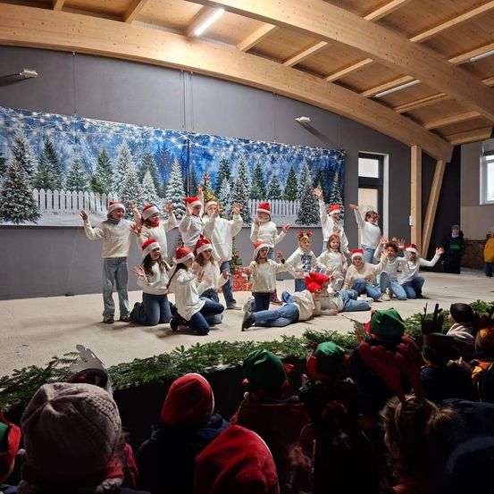 Eine Gruppe von Kindern in festlichen Mützen und Schals tritt auf einer Bühne auf, die mit Weihnachtsbäumen und Grünpflanzen geschmückt ist. Ein Publikum beobachtet von unten.