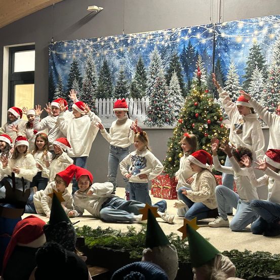 Eine Gruppe von Kindern in Weihnachtsmützen führt ein Weihnachtsspiel auf der Bühne vor einem geschmückten Weihnachtsbaum und Hintergrund auf.