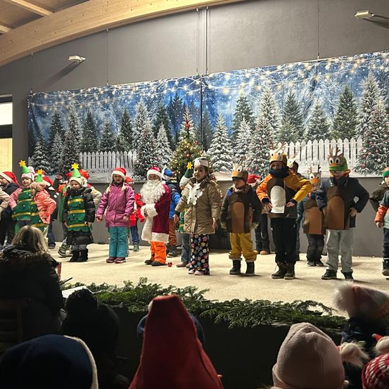Eine Gruppe von Kindern in festlichen Kostümen tritt auf der Bühne vor einem verschneiten Hintergrund auf, mit dem Weihnachtsmann in der Mitte.
