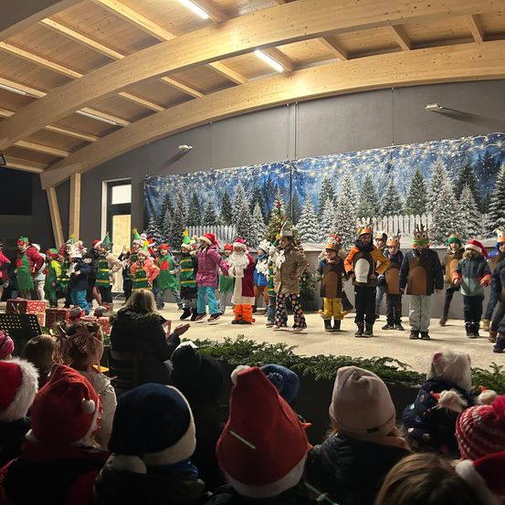 Eine Gruppe von Kindern in Kostümen tritt auf einer Bühne mit einem Weihnachtshintergrund auf, während eine Menge von Menschen in festlichen Mützen von unten zuschaut.