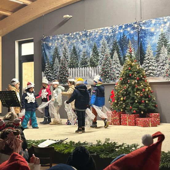 Kinder in Winterkleidung führen auf einer weihnachtlich geschmückten Bühne auf, mit einem Weihnachtsbaum und verpackten Geschenken.
