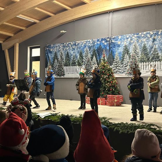 Eine Gruppe von Kindern in Weihnachtskostümen tritt auf einer Bühne auf. Sie haben Rentier-Kopfbedeckungen und Handschuhe. Dahinter steht ein Weihnachtsbaum und eine Kulisse einer verschneiten Landschaft. Ein Publikum schaut zu.