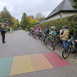 Eine Gruppe von Kindern, alle mit Helmen ausgestattet, stehen mit ihren Fahrrädern an einer Straße in einer Reihe. Ein Polizeibeamter steht an der Seite.