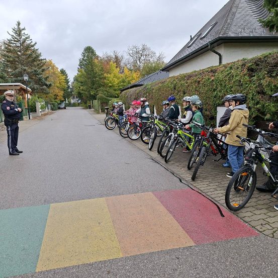 Eine Gruppe von Kindern, alle mit Helmen ausgestattet, stehen mit ihren Fahrrädern an einer Straße in einer Reihe. Ein Polizeibeamter steht an der Seite.