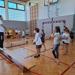 In einer Turnhalle spielen eine Gruppe von Kindern und ein Erwachsener mit Bällen. Sie stehen auf Bänken und halten Bälle, wahrscheinlich spielen sie ein Spiel. Die Turnhalle hat Holzböden, einen Basketballkorb und Glasfenster.